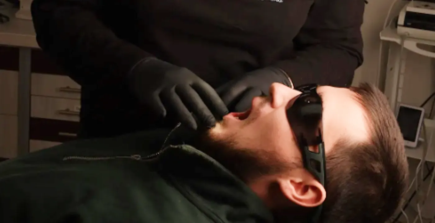 Man wearing dark glasses reclines while a person in black gloves examines his mouth.