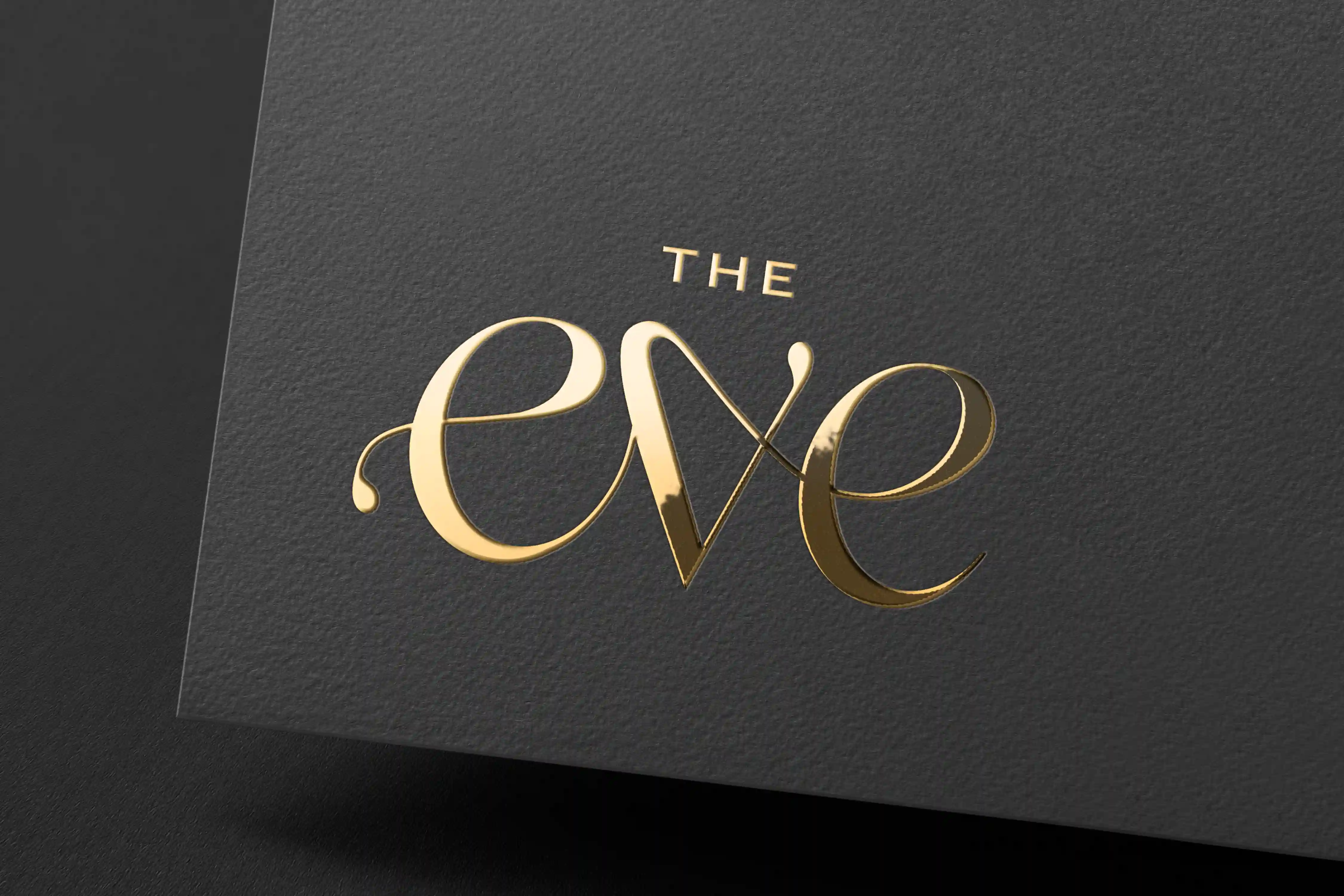 Gold foil embossed logo mockup for The Eve hotel, displaying the brand's elegant script wordmark on a dark charcoal textured card stock.