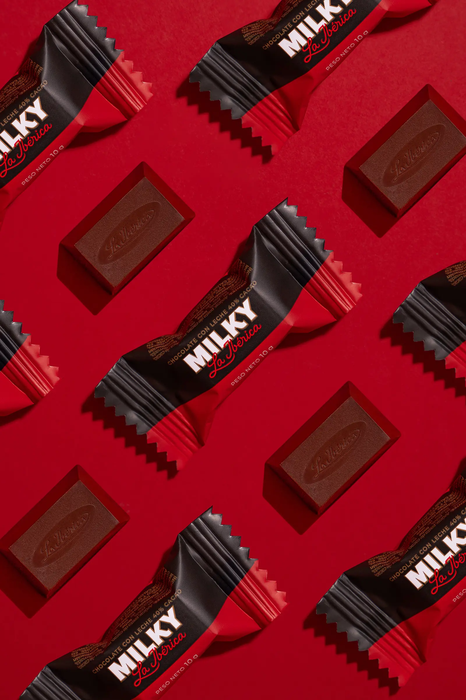 A flat lay of La Ibérica Milky chocolate bars and unwrapped pieces arranged in a repeating diagonal pattern against a bold red background, highlighting the product's black and red packaging design.