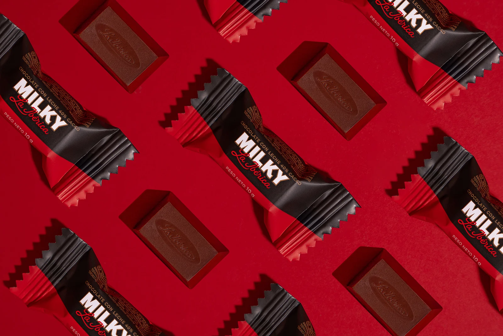 A flat lay of La Ibérica Milky chocolate bars and unwrapped pieces arranged in a repeating diagonal pattern against a bold red background, highlighting the product's black and red packaging design.