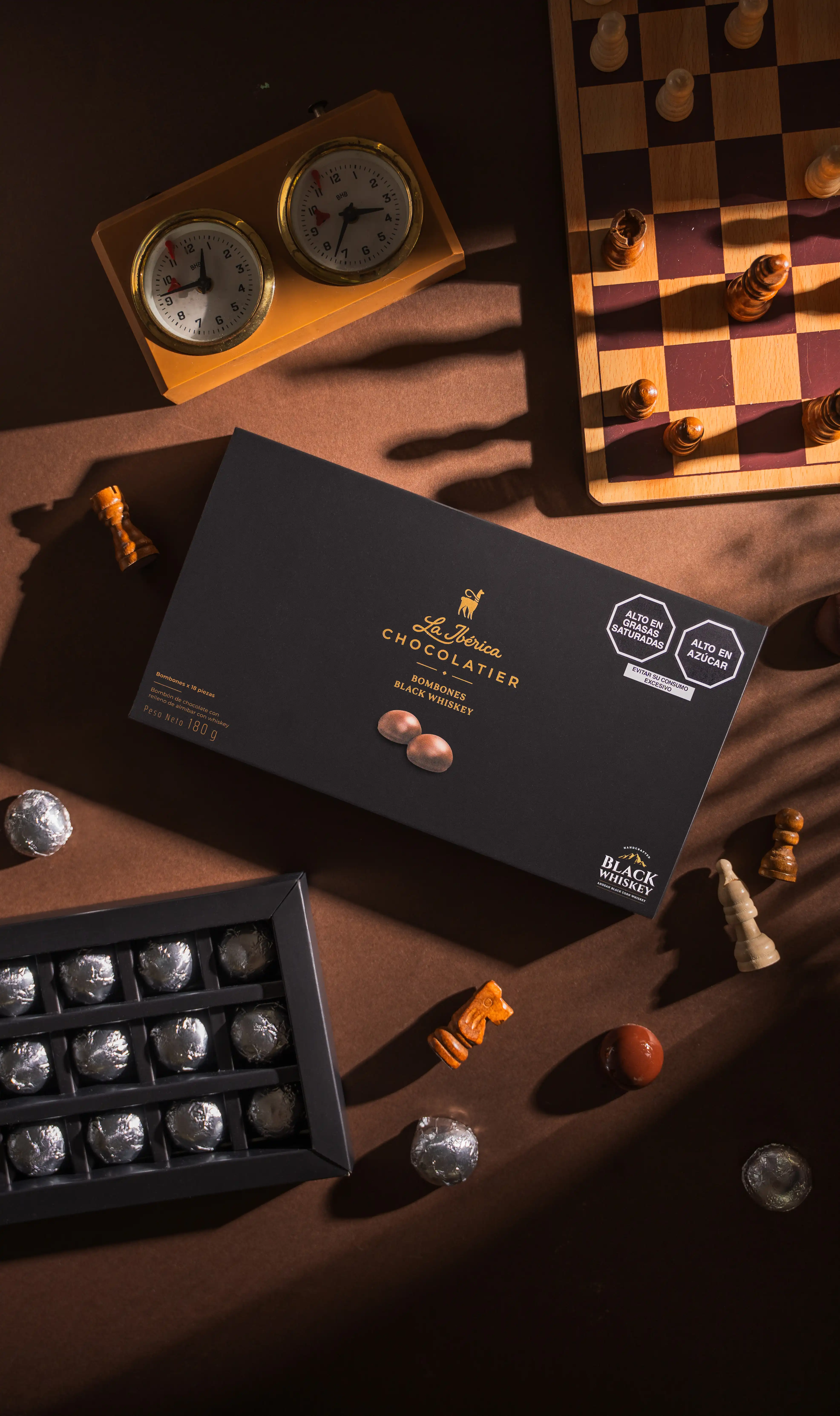 La Ibérica Chocolatier's Bombones Black Whiskey gift box and open tray of silver-wrapped chocolates, styled with a chess board, chess pieces, and a vintage chess clock against a warm brown surface.