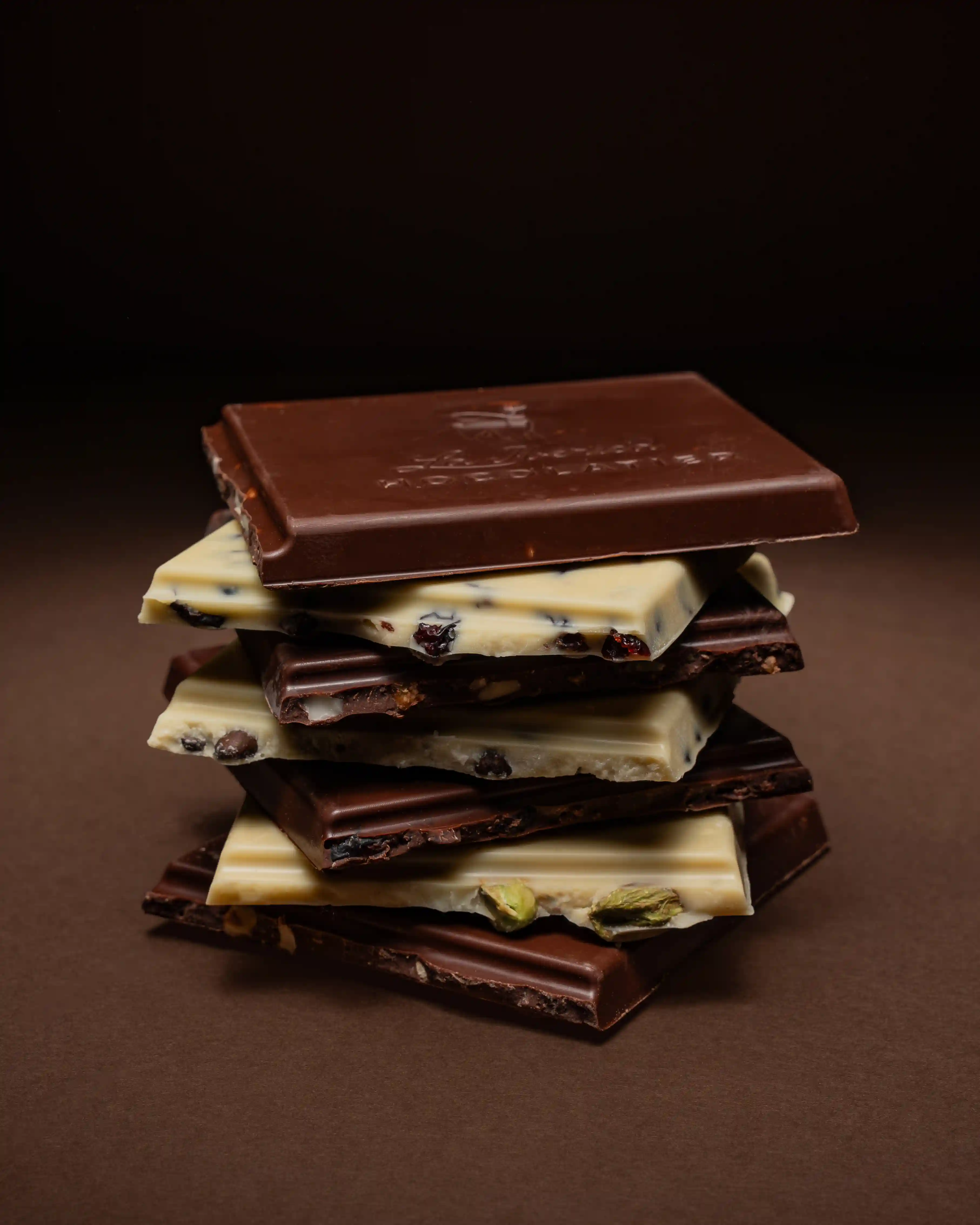 A stack of La Ibérica chocolate tablet pieces in dark, milk, and white varieties with visible inclusions of pistachios, cranberries, and nuts, photographed against a warm brown background.