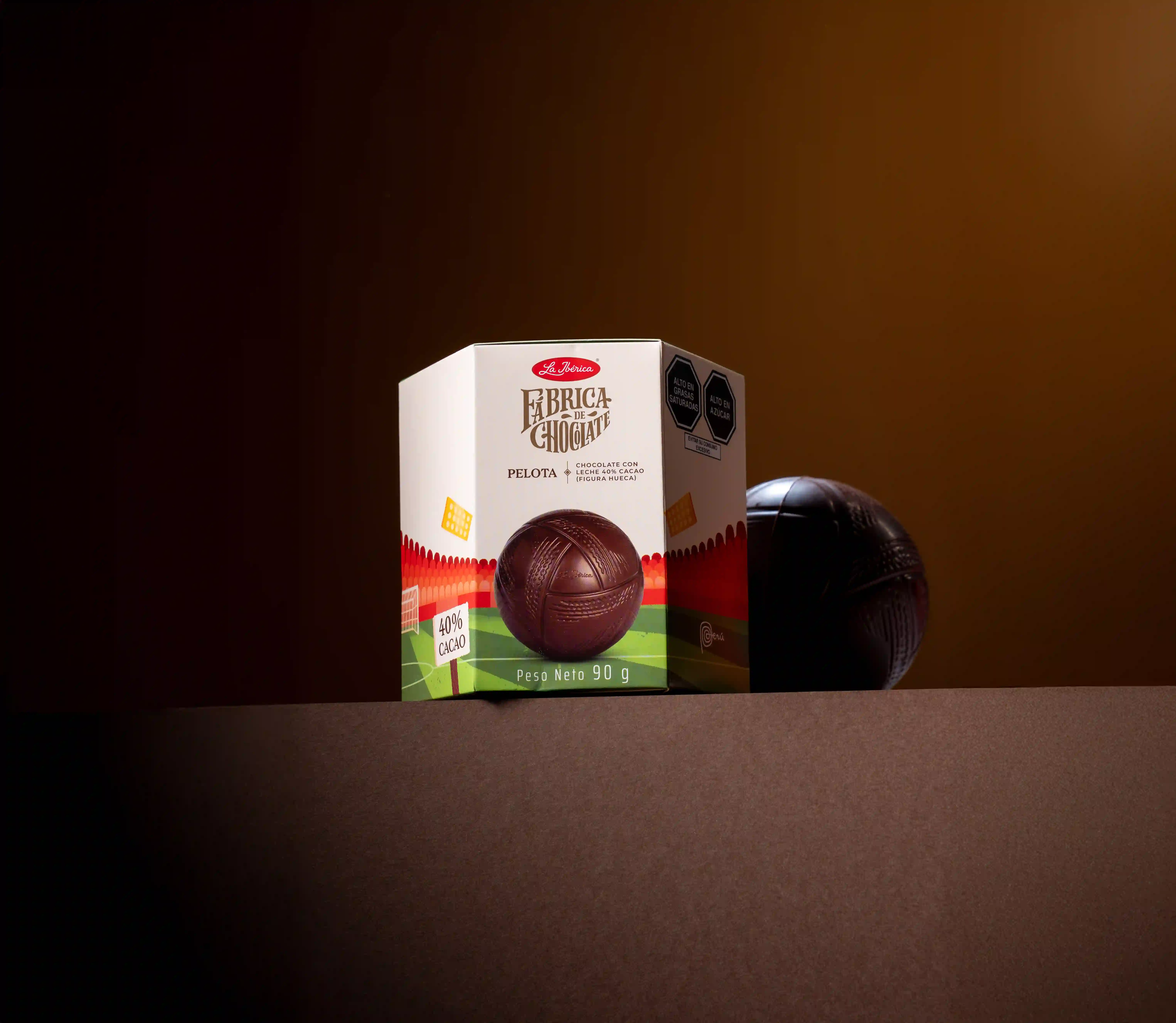 La Ibérica's Fábrica de Chocolate Pelota packaging alongside its hollow milk chocolate soccer ball figure, photographed against a warm dark brown background.