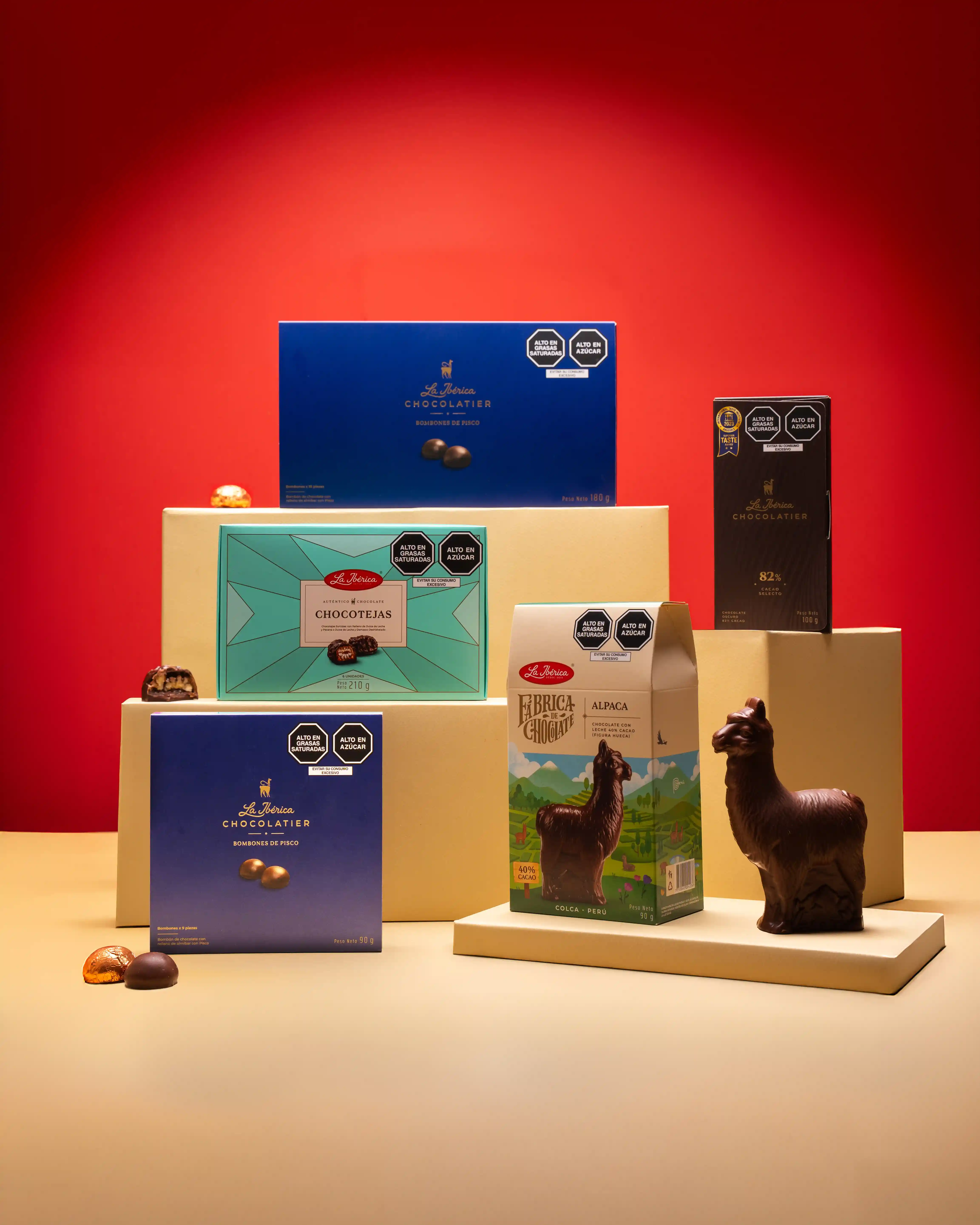 A styled product shot of La Ibérica's full chocolate range arranged on cream pedestals against a red background, featuring the Bombones de Pisco gift boxes, Chocotejas, Fábrica de Chocolate Alpaca carton, 82% dark chocolate bar, and a chocolate alpaca figu