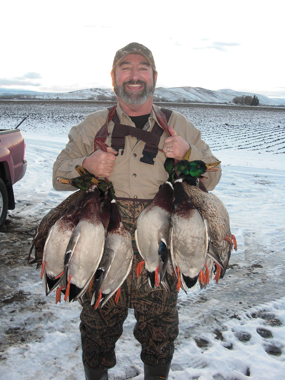 Smiling Mark in camo pants holding eight mallard ducks by straps amid snowy farm fields.
