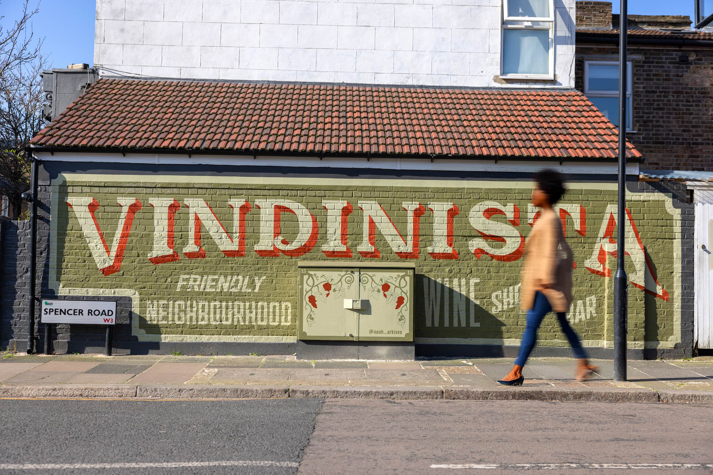 Hand-painted Vindinista wine shop and bar sign on a green brick wall on Spencer Road in Acton, with a woman walking past in motion