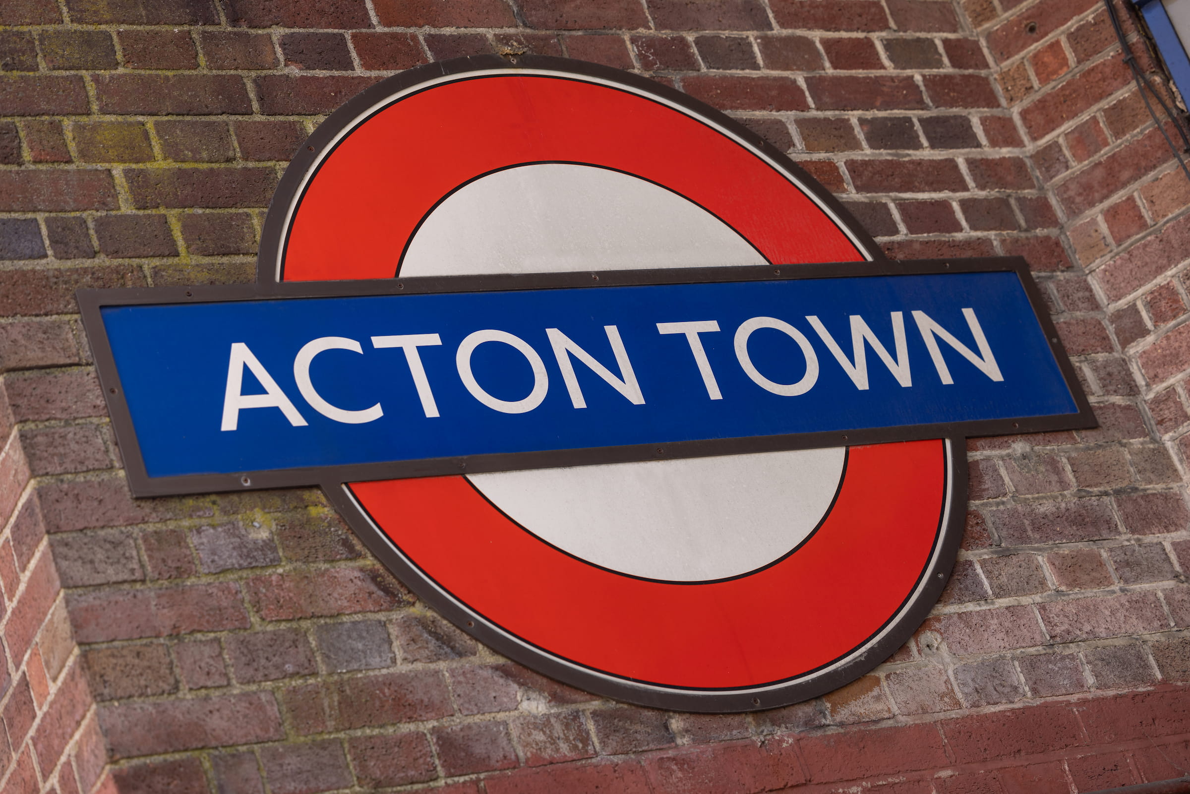 Acton Town London Underground roundel sign in red, white and blue mounted on a weathered red brick wall