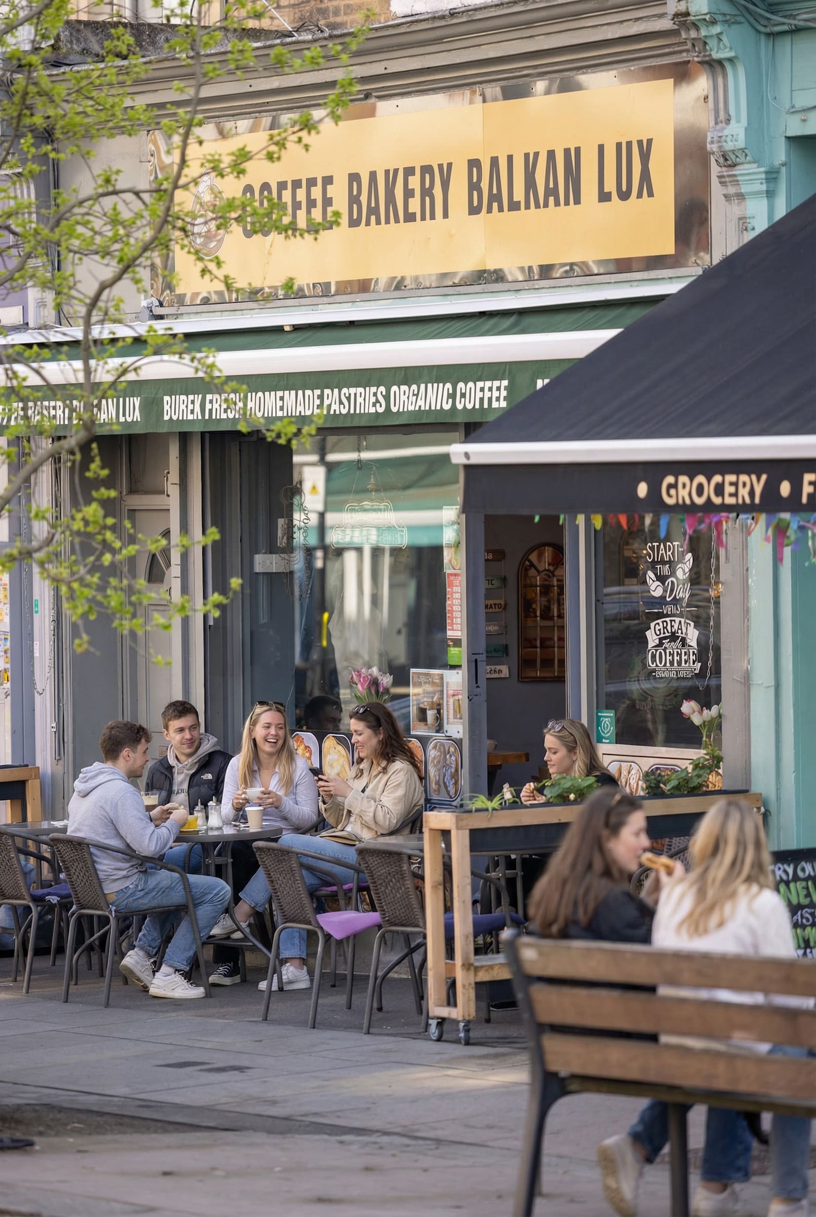 Customers sitting outside Coffee Bakery Balkan Lux in Acton enjoying drinks and pastries on a sunny day, with a yellow signboard above