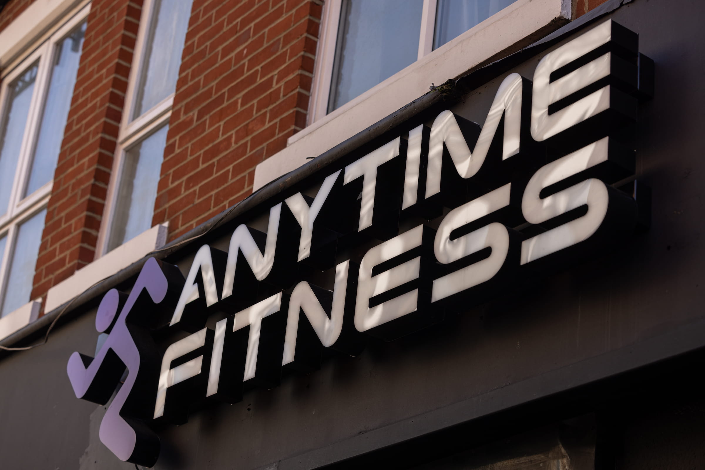 Anytime Fitness illuminated signage with white block letters and a purple running figure logo on a red brick building in Acton