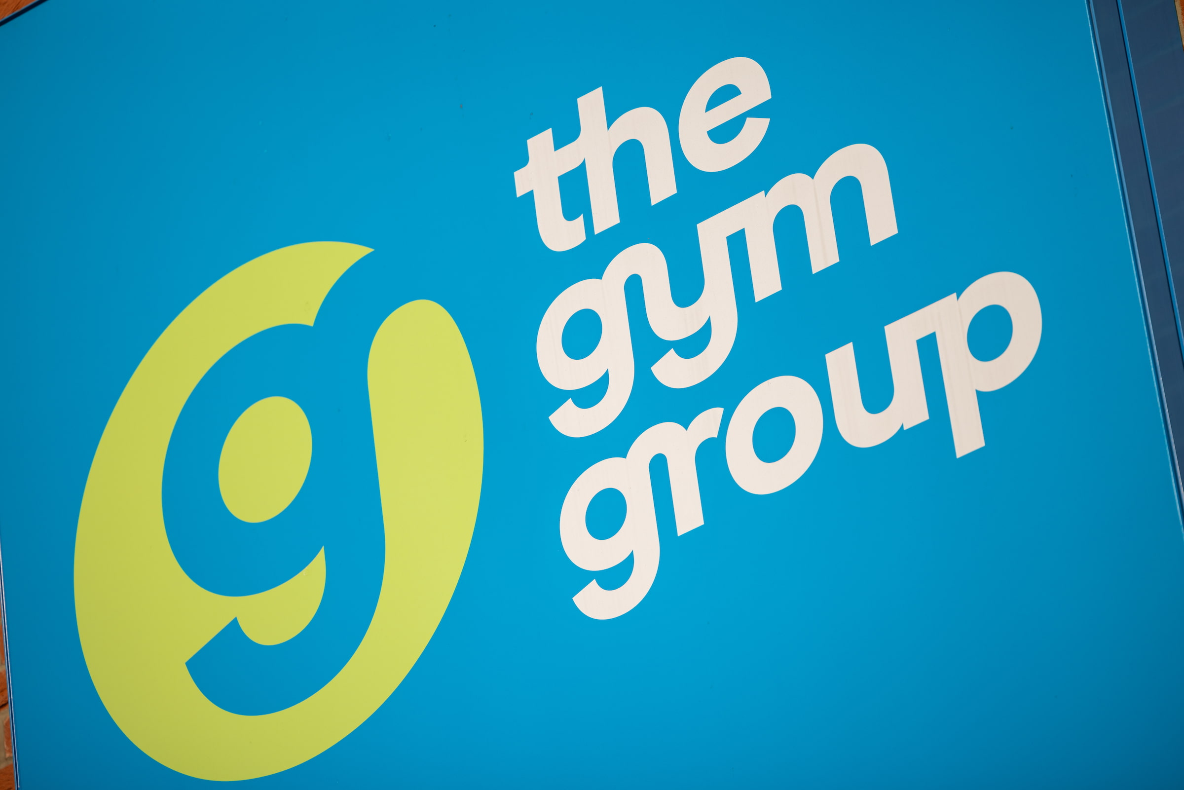 The Gym Group logo featuring a lime green "g" symbol and white lowercase text on a bright blue background