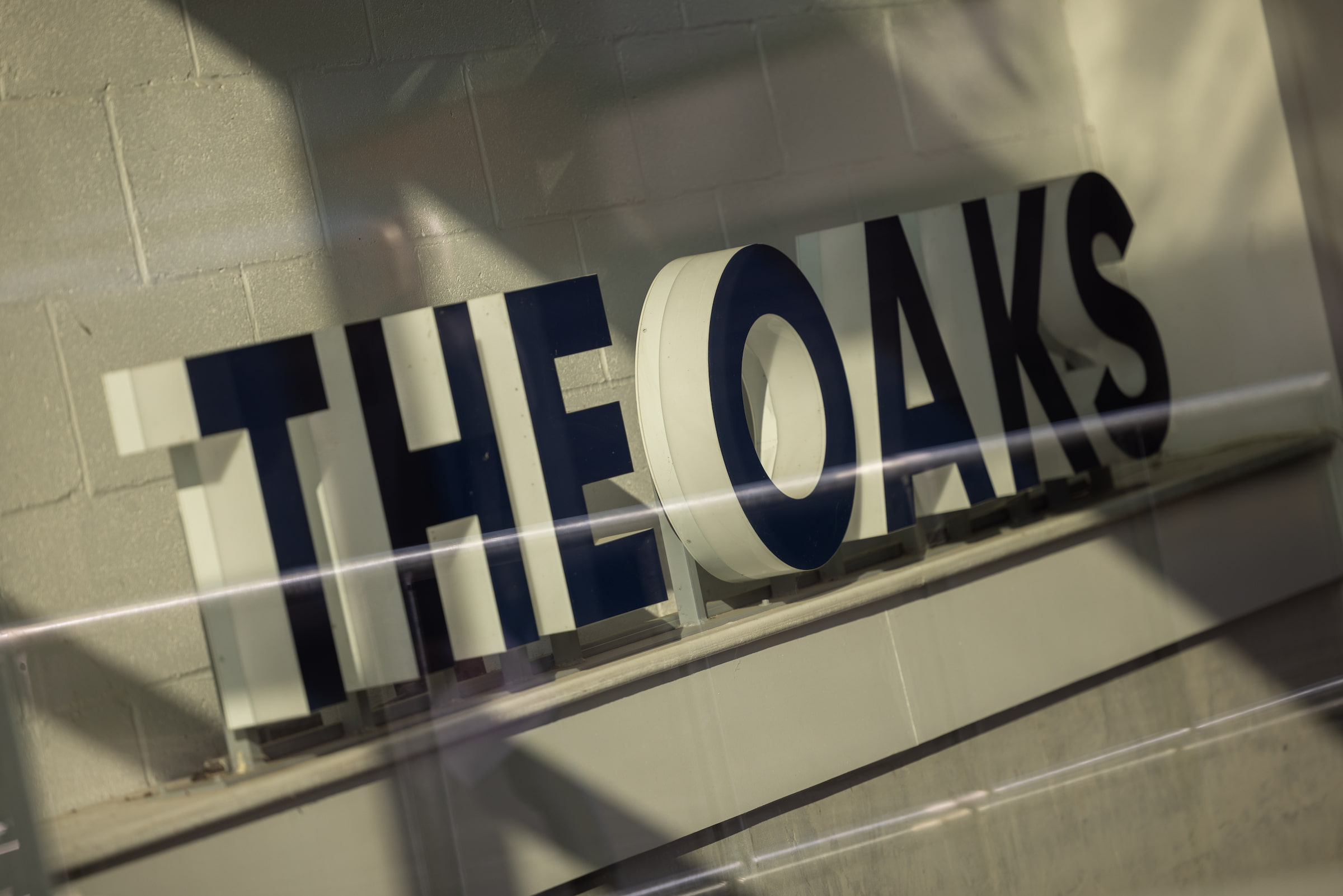 Close-up of The Oaks signage with bold navy blue lettering mounted on a white brick wall, viewed through a glass railing in Acton, London