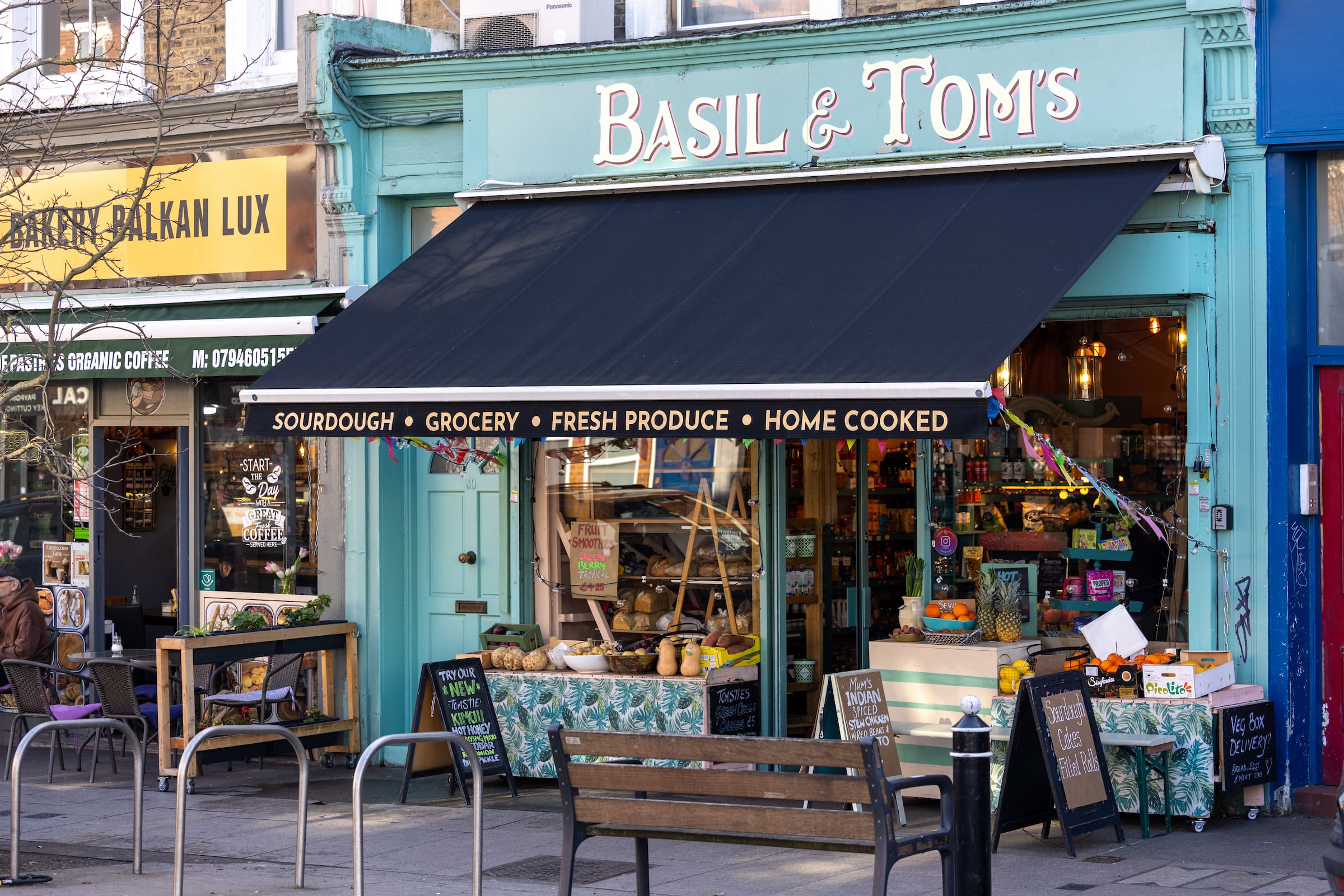 Basil and Tom's shopfront with a turquoise facade, navy awning advertising sourdough, grocery, fresh produce and home cooked food, next to Coffee Bakery Balkan Lux