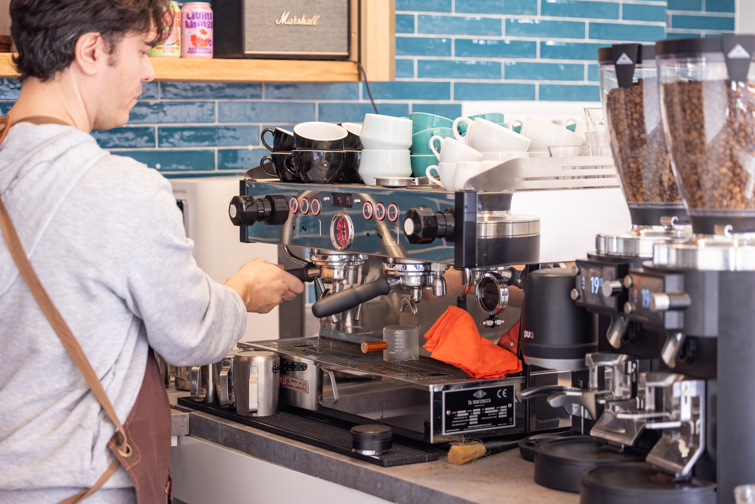 Barista in a grey jumper and leather apron operating a La Marzocco espresso machine in front of a teal tiled wall with stacked coffee cups in an Acton café
