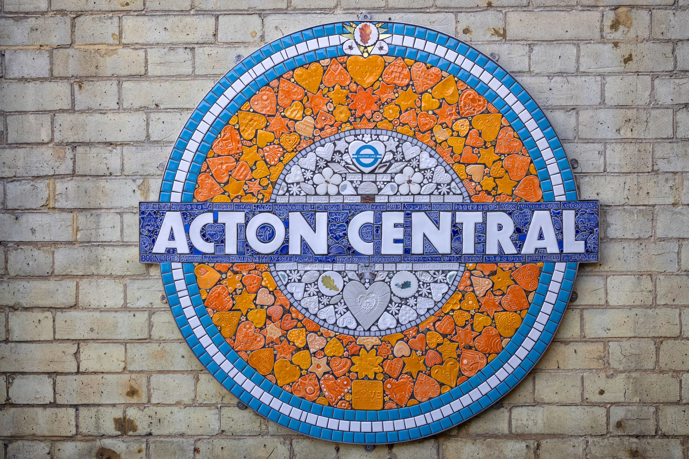 Acton Central station roundel sign in mosaic tiles featuring orange hearts, stars and community symbols mounted on a yellow brick wall