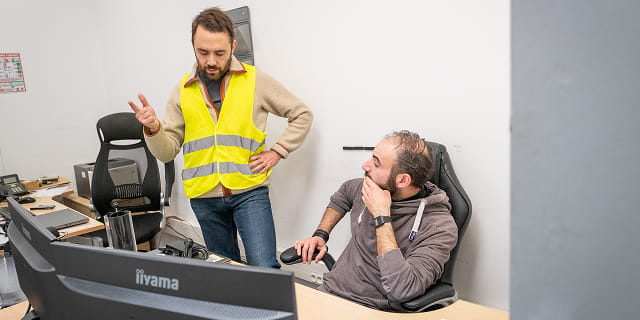 Deux hommes discutent dans un bureau, l'un debout avec un gilet jaune, l'autre assis devant un écran d'ordinateur.