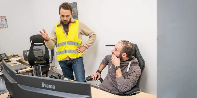 Deux hommes discutent dans un bureau, l'un debout avec un gilet jaune, l'autre assis devant un écran d'ordinateur.