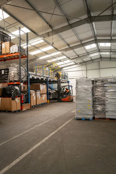 Entrepôt avec palettes en carton et plastique, chariot élévateur et rayonnages sous un toit métallique.