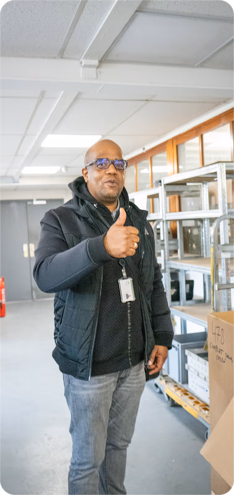 Man wearing glasses and black jacket giving a thumbs-up gesture in a storage room.