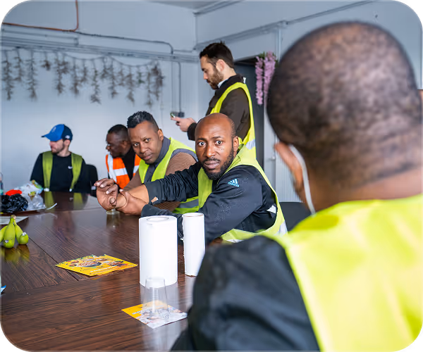 Workers in safety vests seated around a table, some looking toward the camera during a meeting.