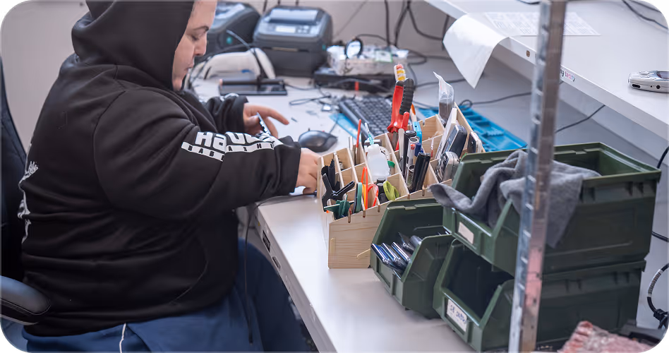 Person in black hoodie working at desk with organized tools and electronic devices in bins.