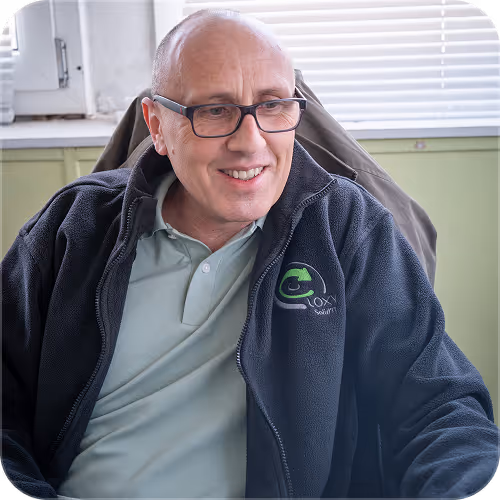 Smiling bald man with glasses wearing a black jacket indoors near a window with blinds.