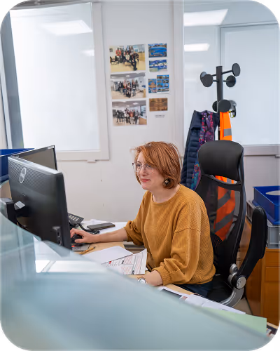 Woman with red hair and glasses working at a desk on a computer in an office setting.