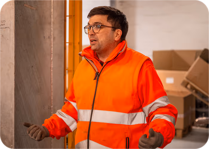 Man in orange reflective safety jacket and gloves talking in an industrial or warehouse setting.