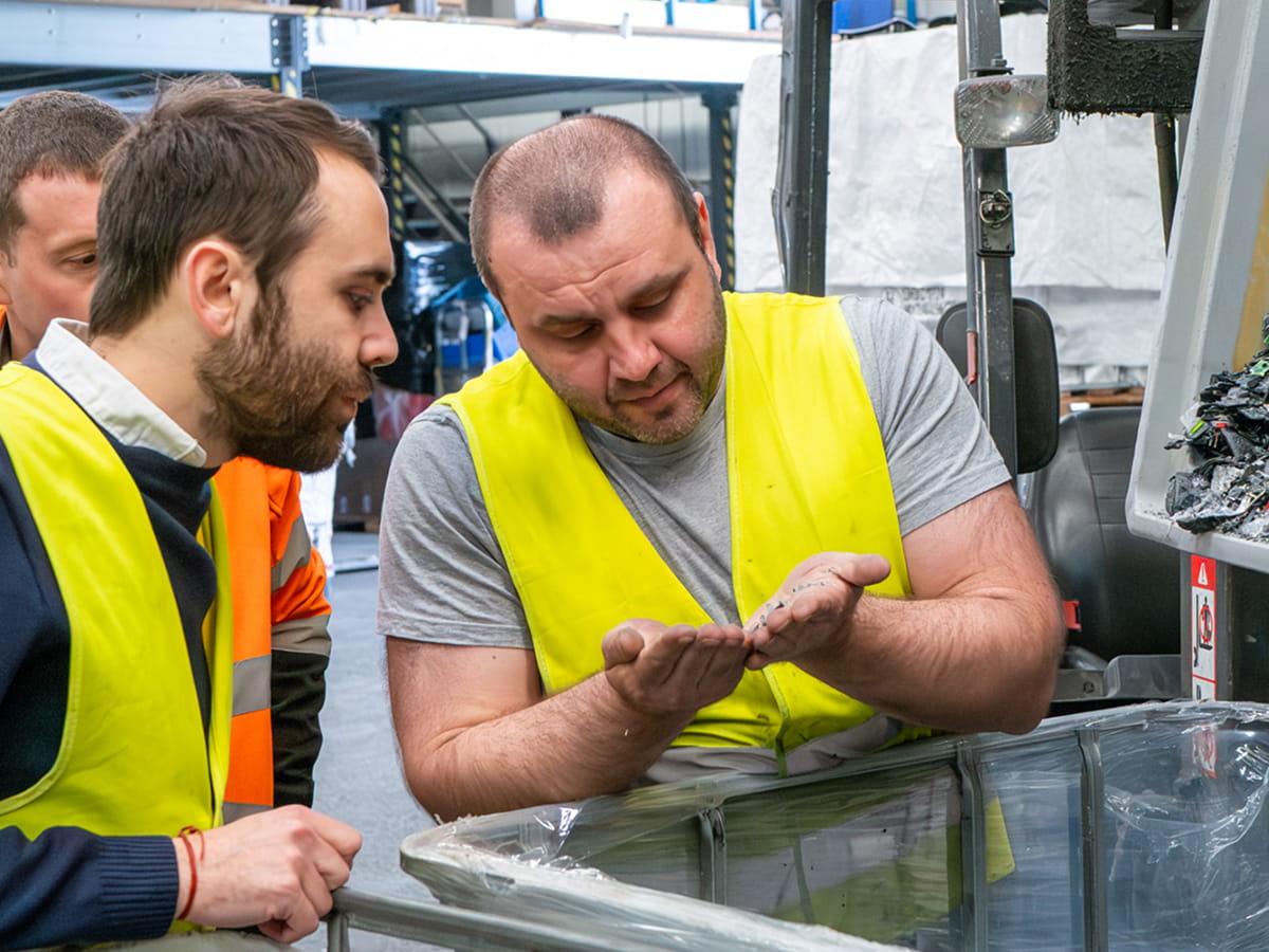 Trois travailleurs portant des gilets de sécurité observent des granulés recyclés dans la main de l'un d'eux.
