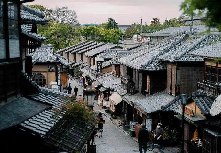 Zijaanzicht van een sfeervolle, hellende straat in een historische Japanse wijk met traditionele houten huizen, dongrijze dakpannen en rustig wandelende buurtbewoners.