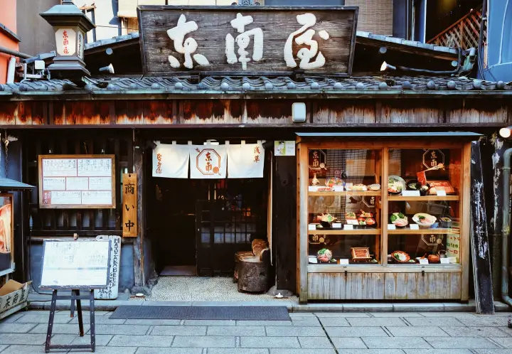 De houten voorgevel van een authentiek Japans restaurant met een grote houten naambord boven de ingang, een traditioneel noren-gordijn en een glazen vitrine vol voorbeelden van gezonde Japanse gerechten.