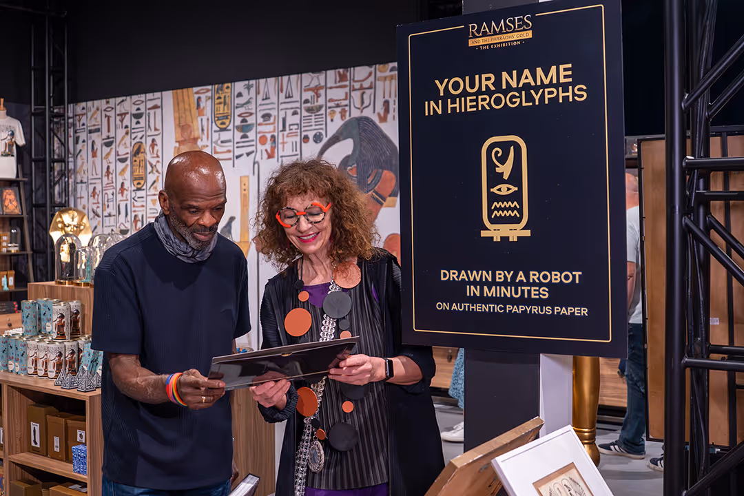 Two people looking at framed papyrus art near a sign about names in hieroglyphs drawn by a robot.