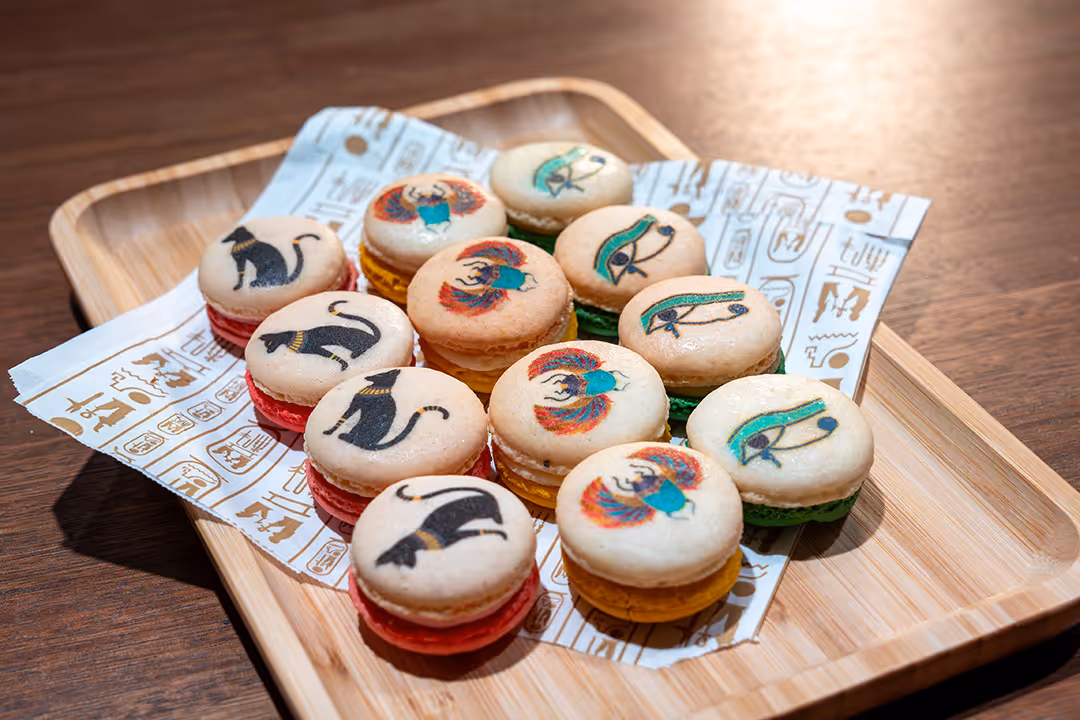 Macarons decorated with Egyptian-style art of cats, birds, and eyes on a wooden tray with patterned paper.