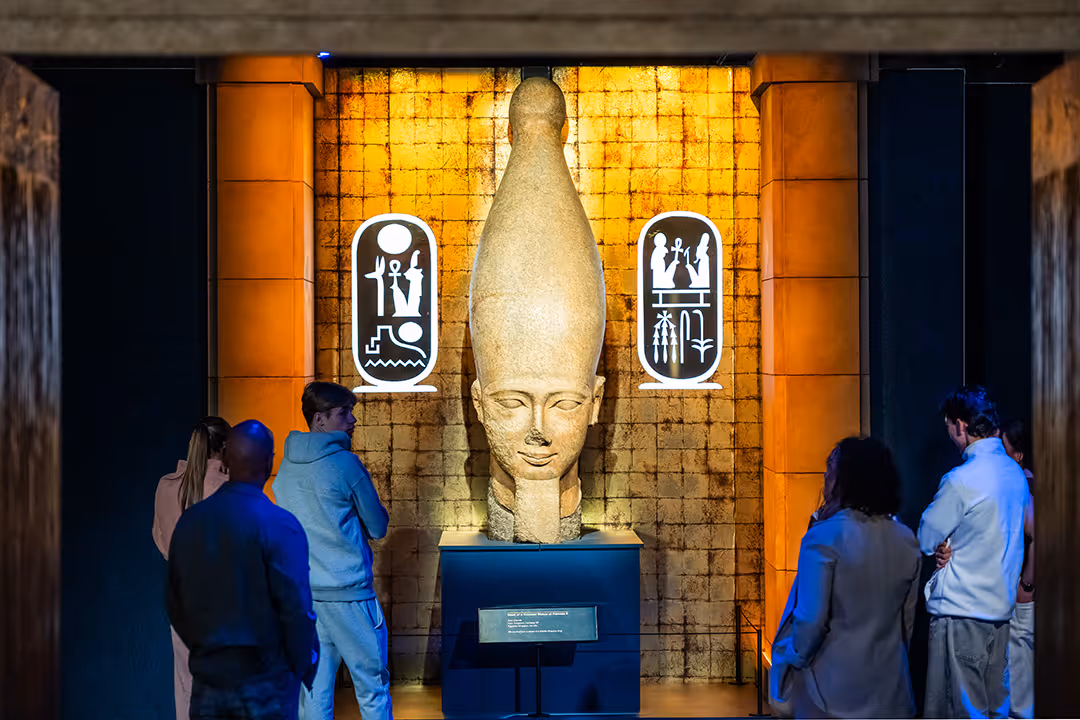 Large ancient Egyptian statue head with tall headdress displayed with visitors observing.