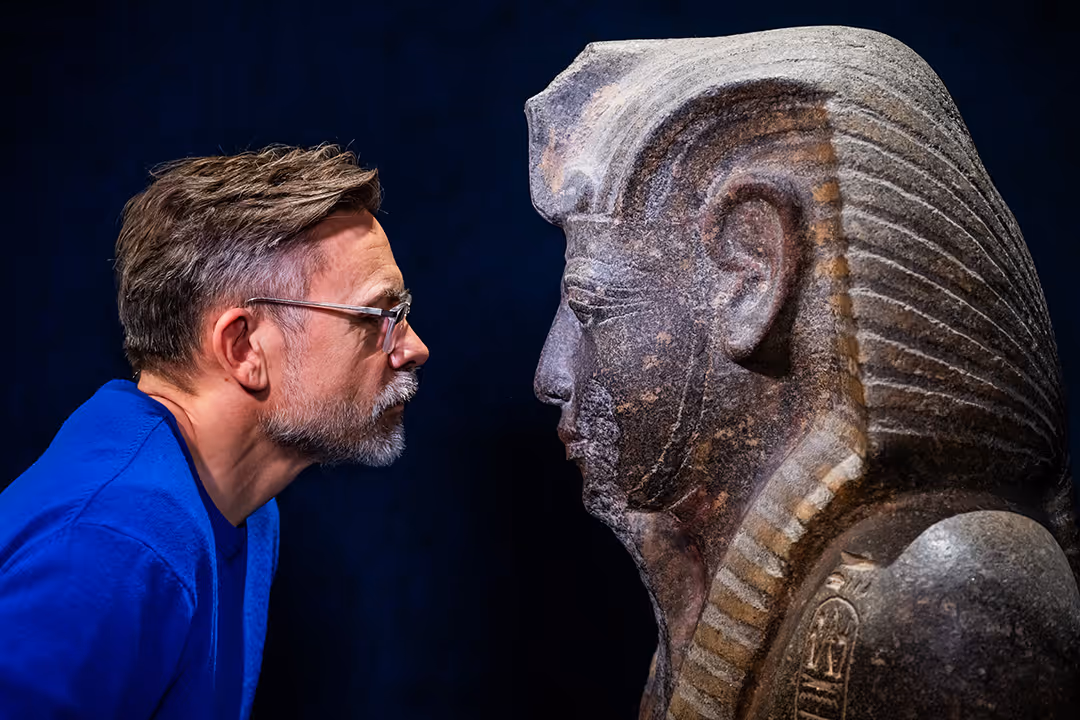 Man wearing glasses closely examines the profile of an ancient Egyptian statue against a dark background.