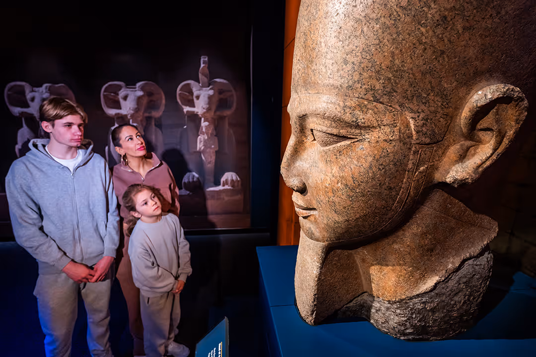 A woman and two children observe a large ancient Egyptian stone head sculpture in Ramses The Exhibition.