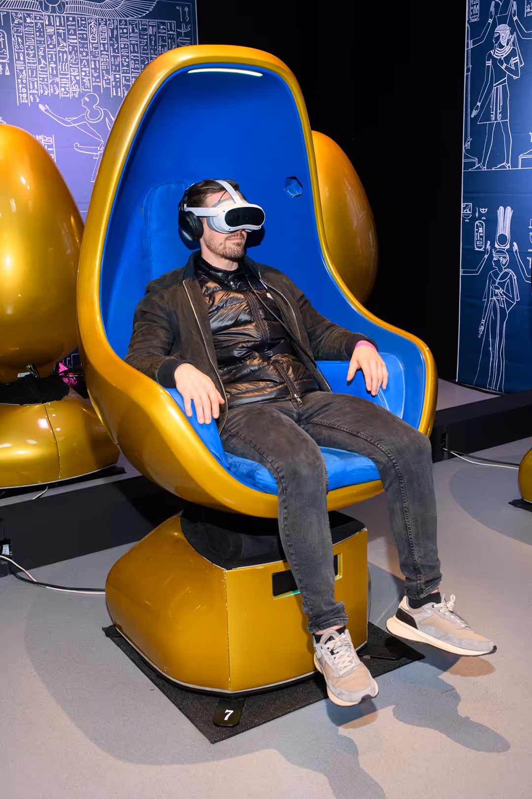 Man wearing VR headset sits in a futuristic yellow and blue gaming chair with Egyptian decor.