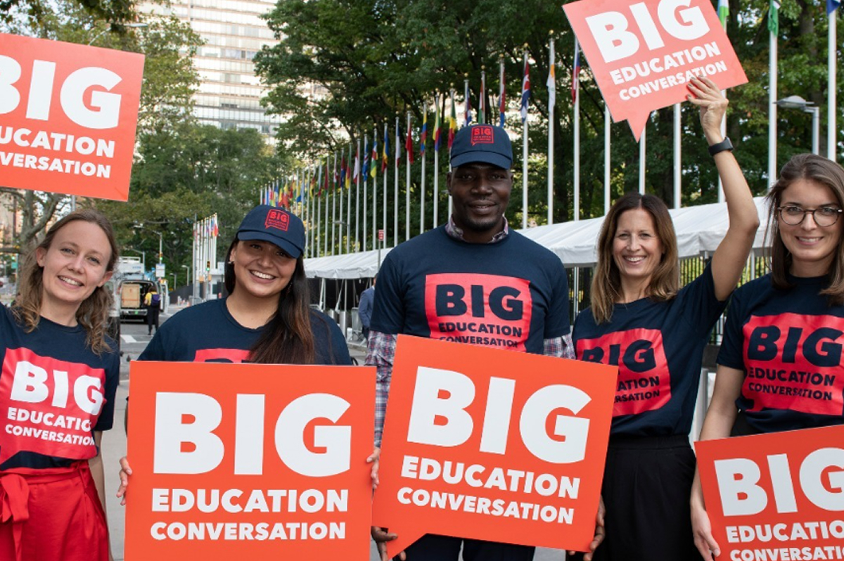 Five smiling Big Change campaigners stand outdoors in front of international flagpoles, wearing dark "BIG EDUCATION CONVERSATION" t-shirts and caps and holding orange placards with the same white text.