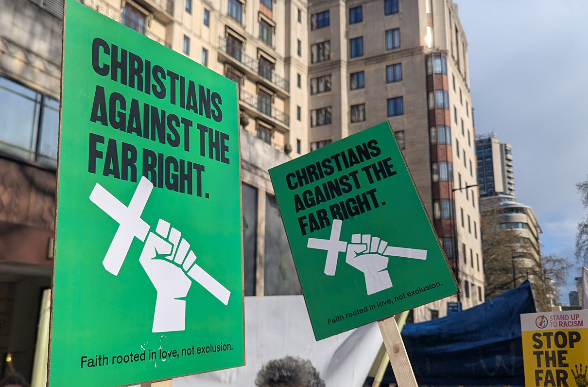 Two large green placards held aloft at a city-centre protest in front of a tall office building, each featuring a white raised fist gripping a cross and the slogan "Christians Against The Far Right — Faith rooted in love, not exclusion." A yellow "Stand Up To Racism — Stop The Far…" placard is partially visible on the right.