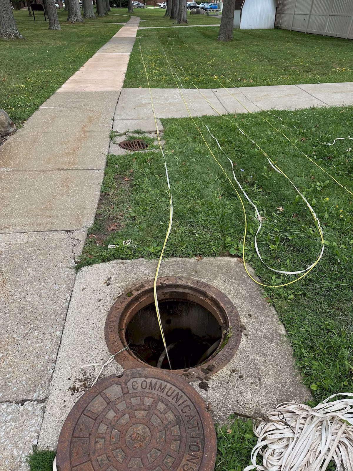 Open communications manhole with fiber cable and pulling tape routed across a multifamily property lawn.