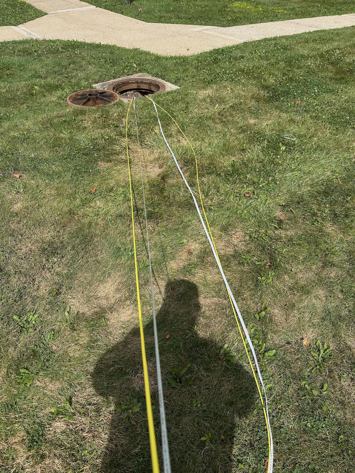 Yellow fiber cable and white pulling tape laid across grass toward an open underground communications manhole.