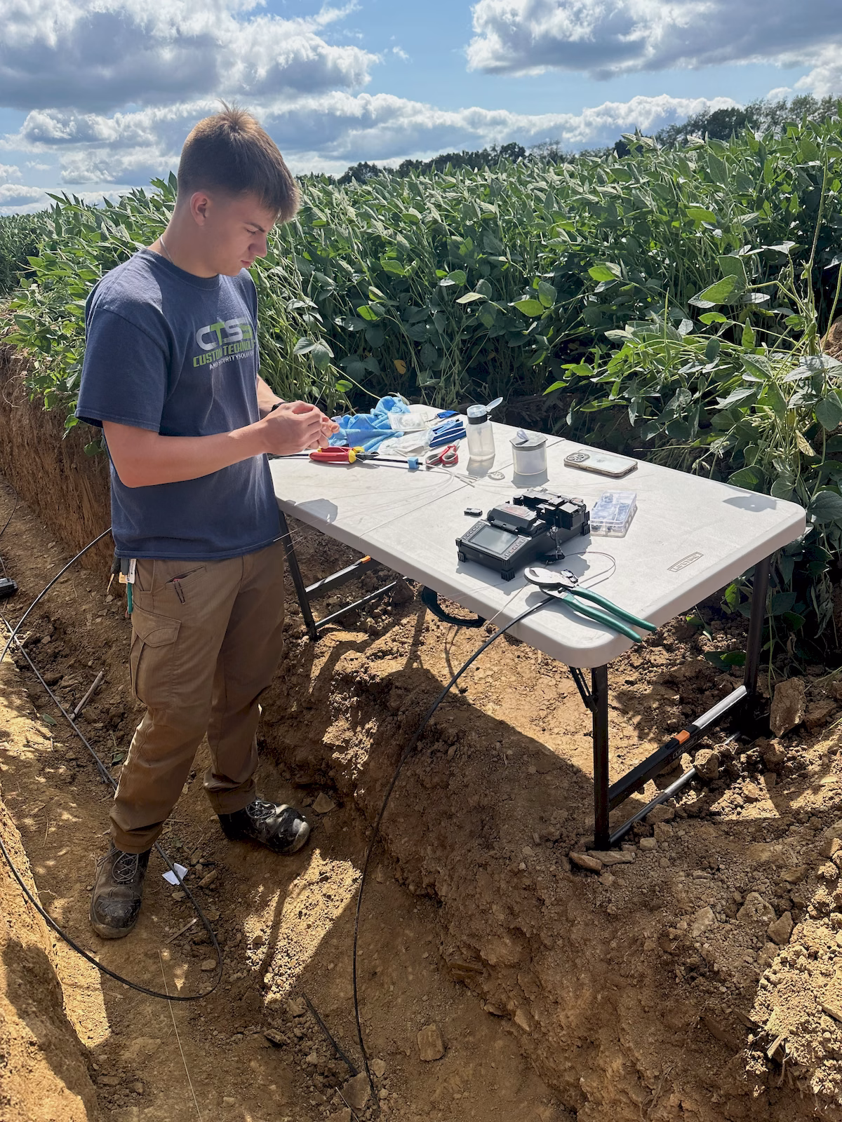 CTSS technician fusion-splicing underground fiber-optic cable beside an excavated field trench.