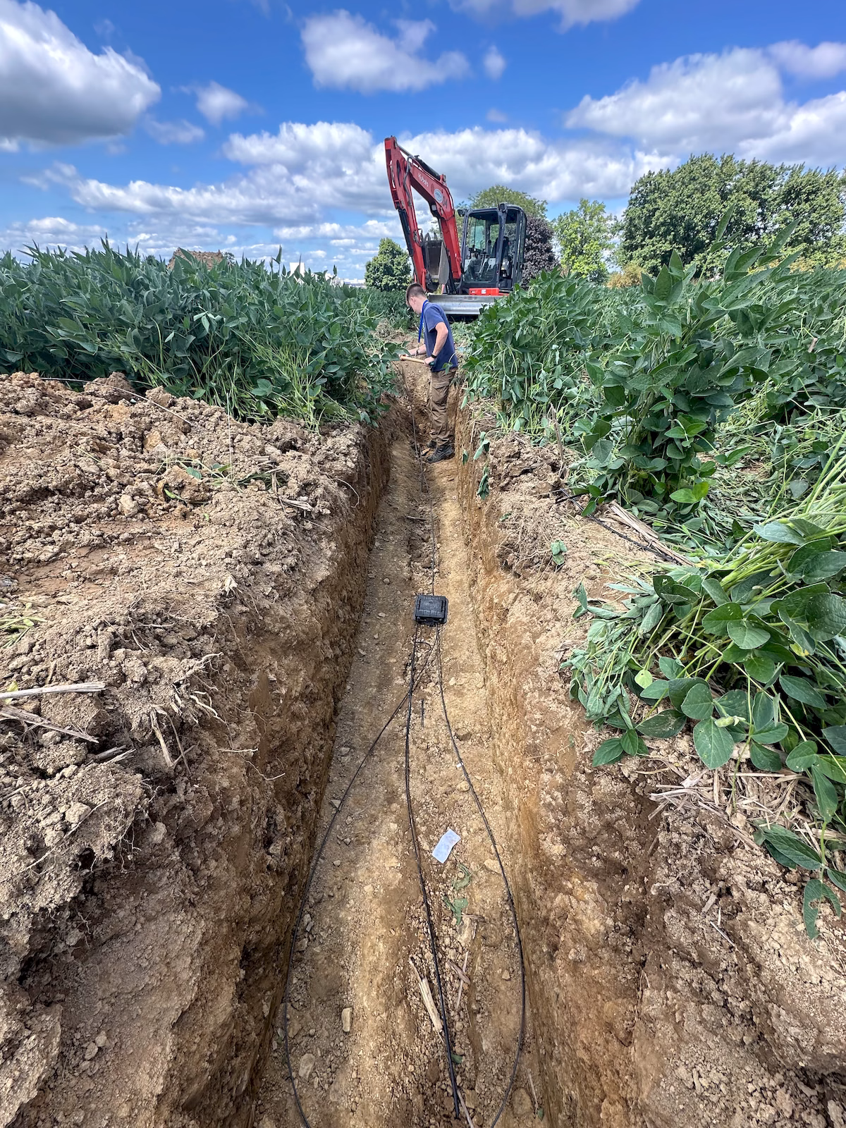 Excavated underground fiber route crossing a field with a technician and compact excavator at the repair site.
