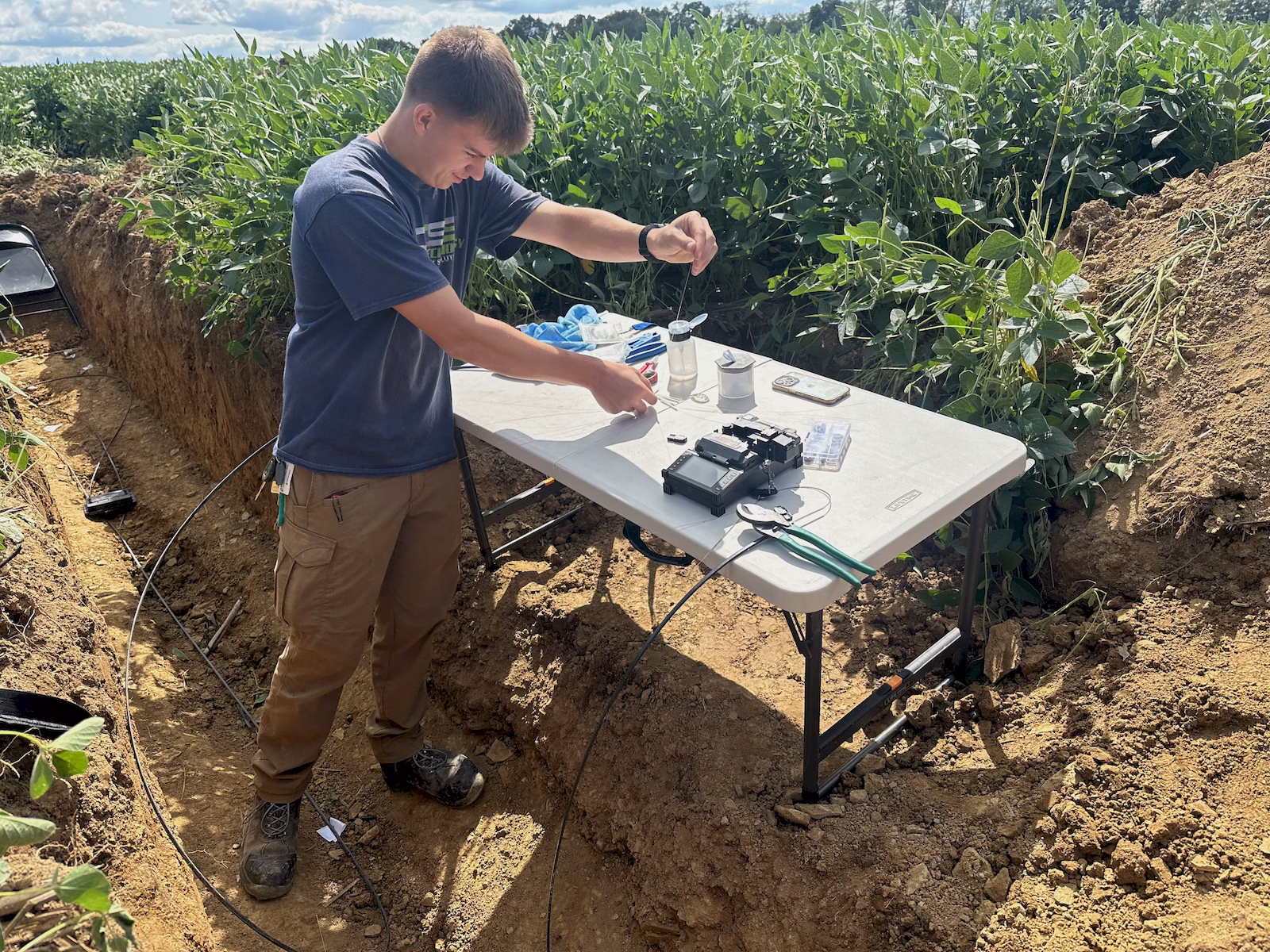 Fiber technician preparing optical fibers beside a fusion splicer at an outdoor repair site.