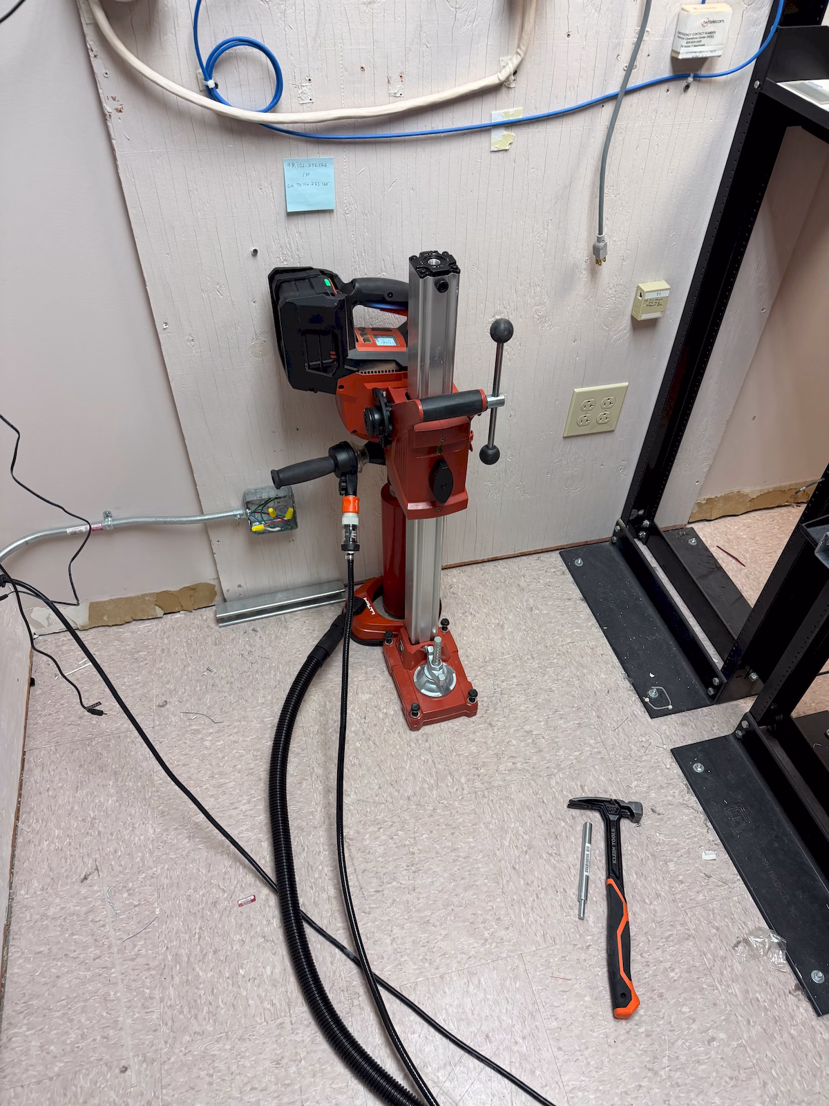 Anchored concrete core-drilling rig positioned beside communications equipment inside a commercial facility.