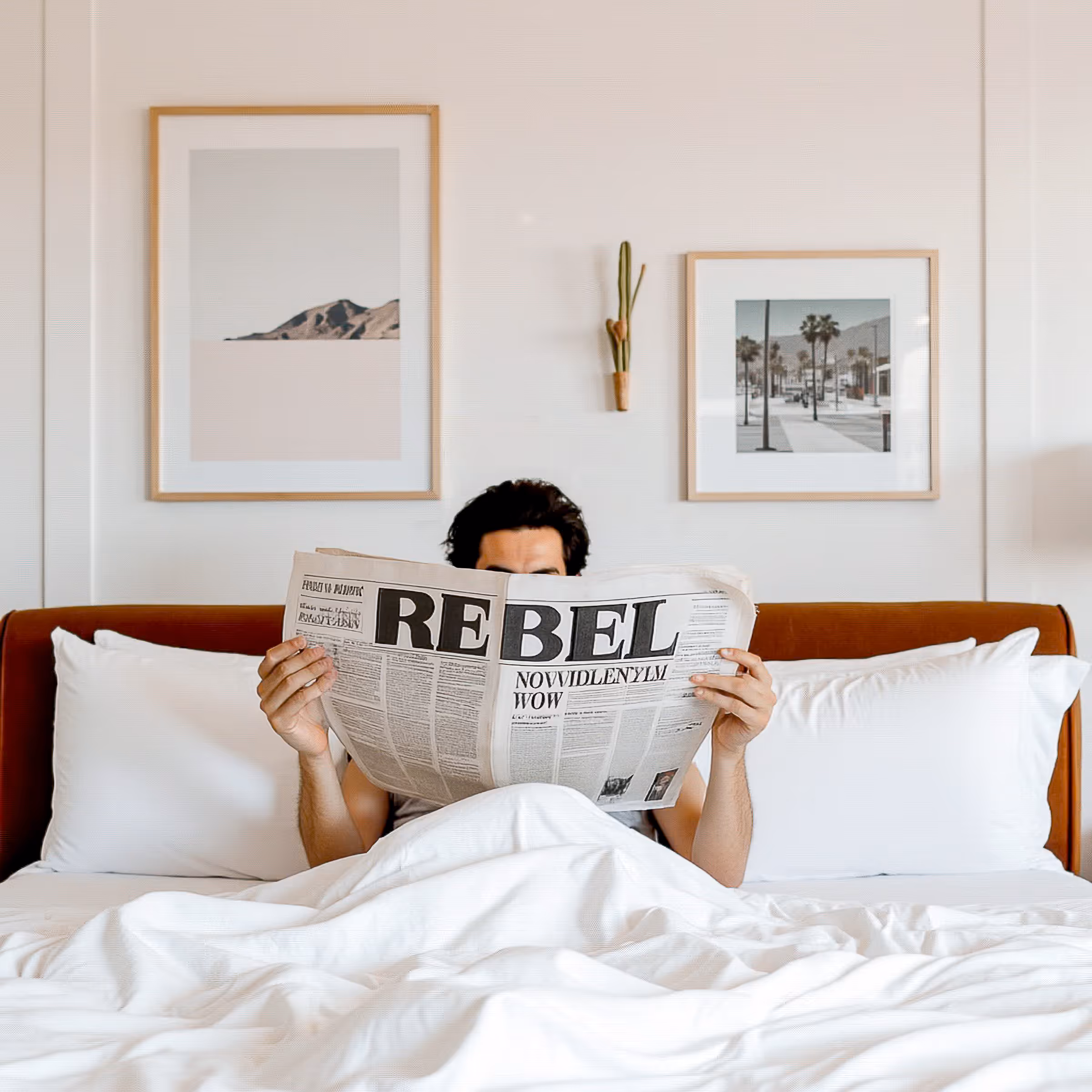 Person sitting in white bed reading a newspaper titled REBEL with two framed photos on the wall.
