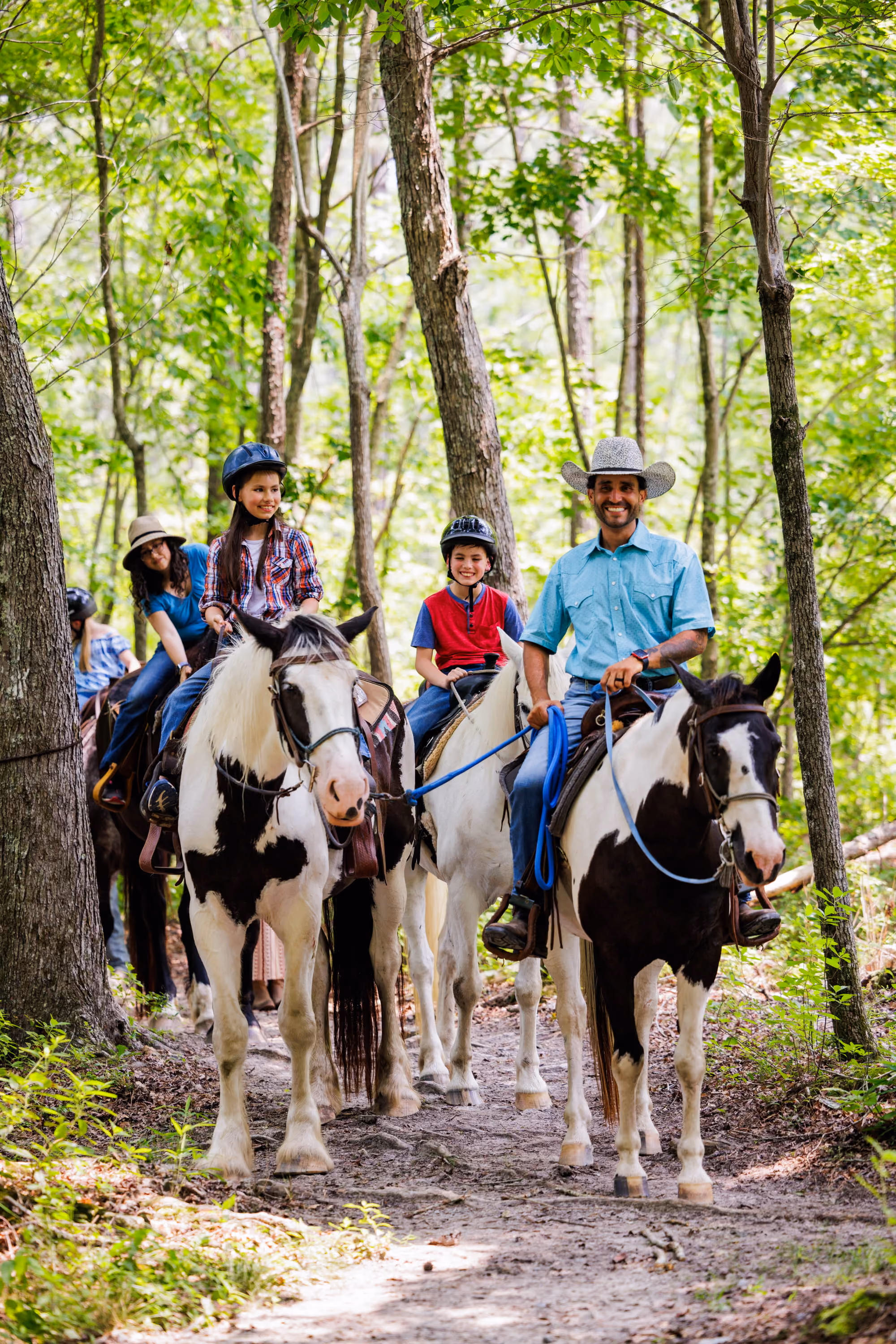 A smiling guide in a cowboy hat and children in riding helmets ride paint horses along a sunlit forest trail.