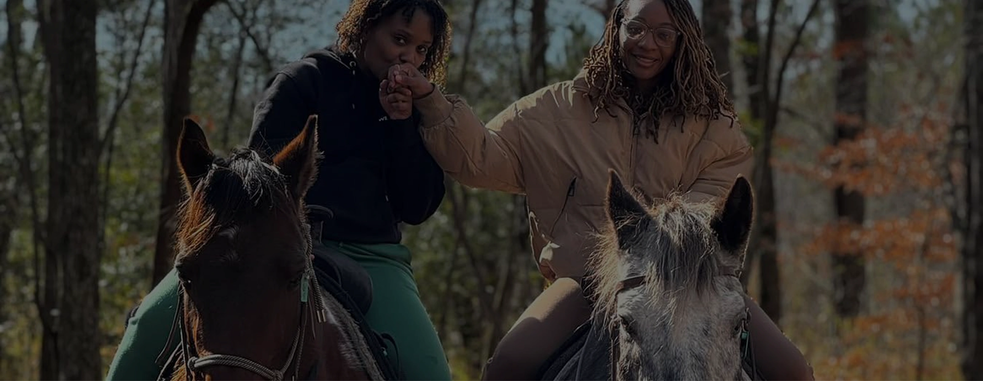 Two women on horseback hold hands, one kissing the other's hand in a wooded outdoor setting.