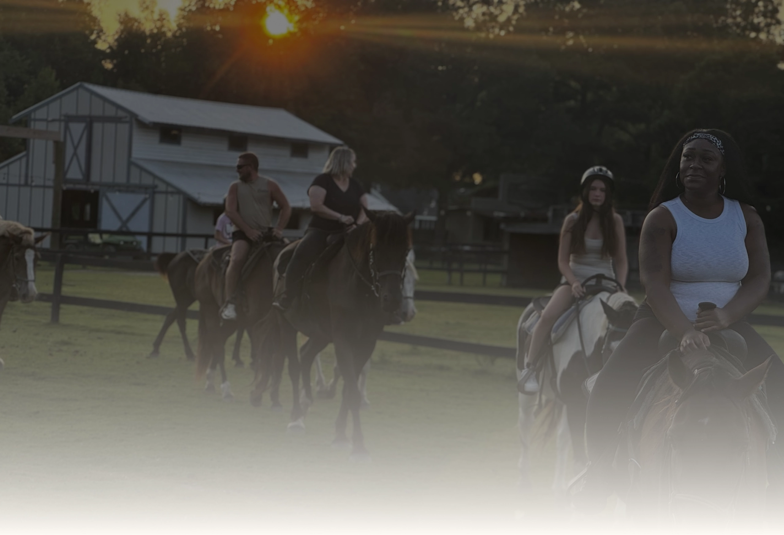 Riders on horseback cross a grassy field at sunset, with the ranch barn behind them.