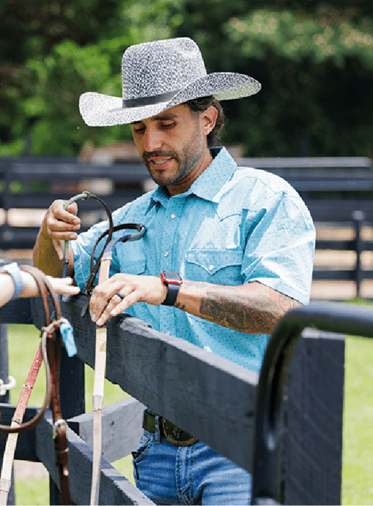 A ranch hand in a cowboy hat adjusts leather reins over a black wooden fence.