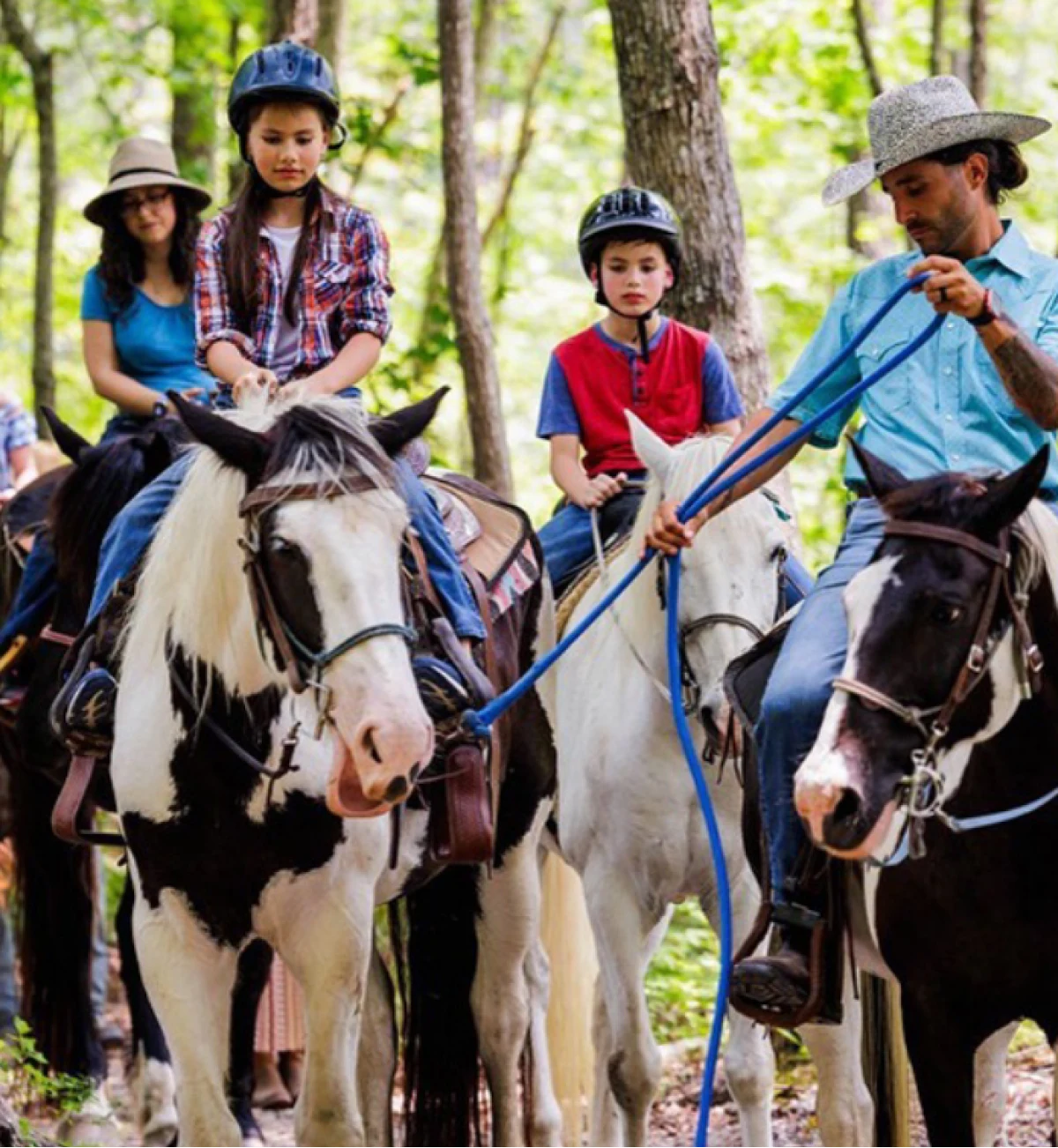 A family group rides horses along a sunlit wooded trail, led by a girl on a pinto and a guide in a cowboy hat.